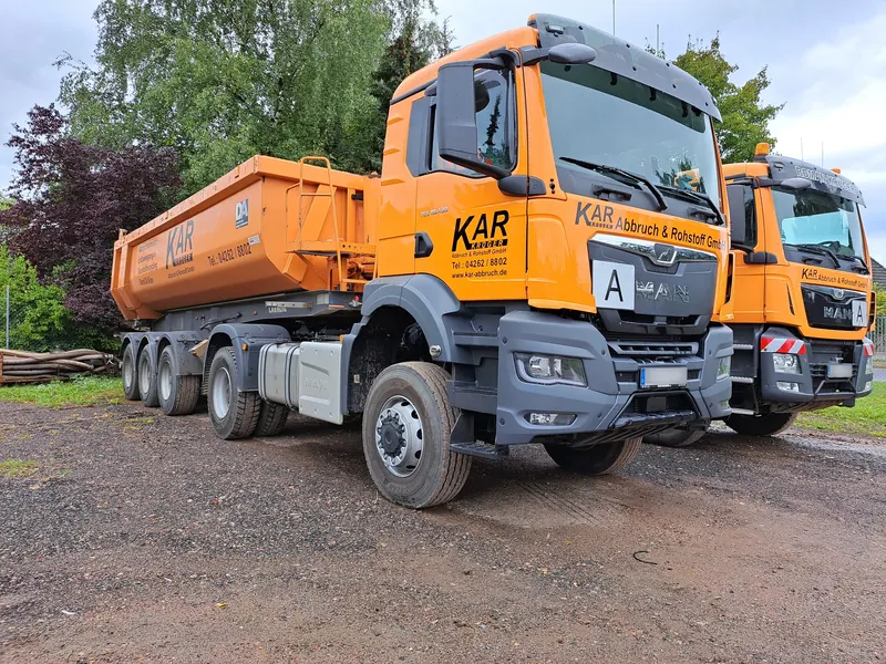 Orangefarbene Sattelzugmaschine mit Kippauflieger und Firmenbeschriftung, daneben ein zweiter Lkw im gleichen Design