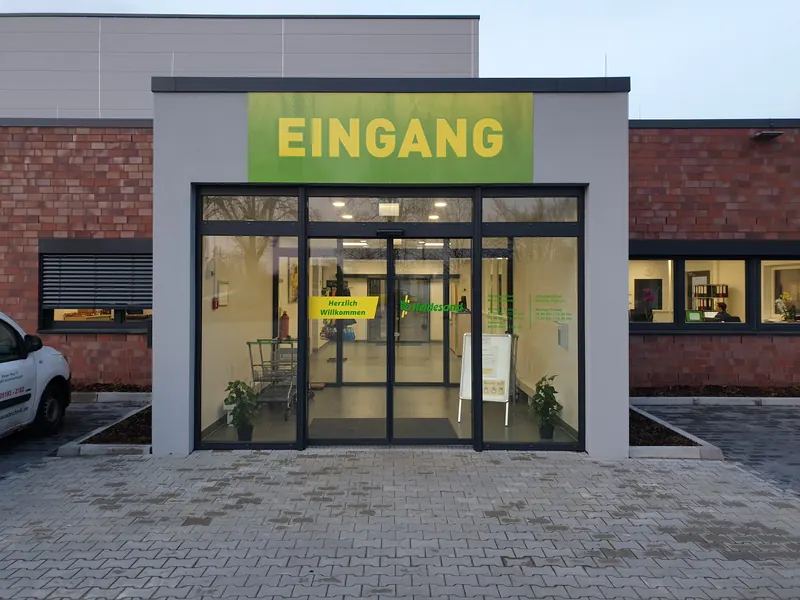 Eingang eines Marktes mit großem Eingangsschild und Folienschrift auf den Glastüren